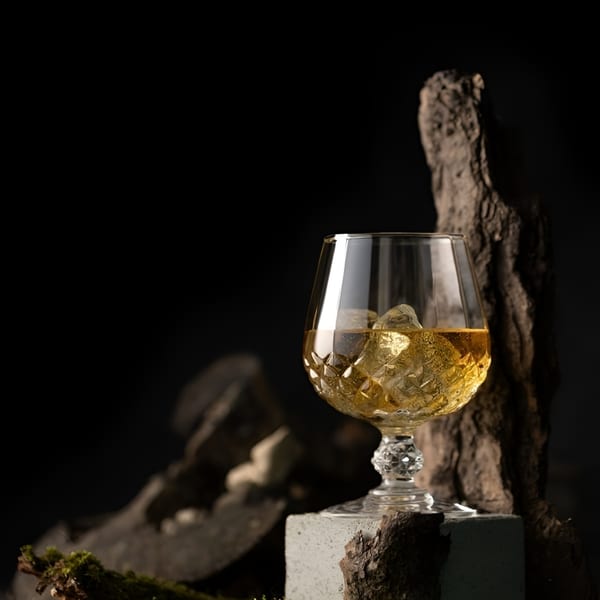 Whiskey with ice in a cut crystal glass, surrounded by dark wood and moss.