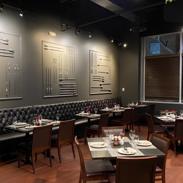 A dimly lit restaurant interior with dark tufted booths, wooden chairs, and set tables.