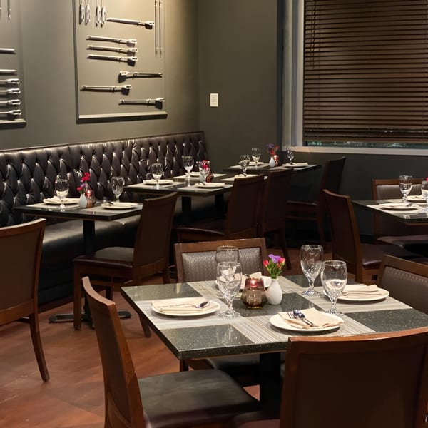 Restaurant dining room with set tables, dark tufted booths, and wall decor featuring decorative metal utensils.