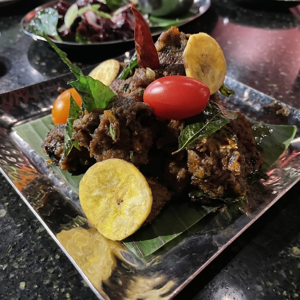 Close-up of a dark, savory dish garnished with cherry tomatoes, banana chips, and a red chili.