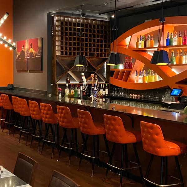 A modern bar with orange bar stools, a dark countertop, and an orange-lit liquor display.
