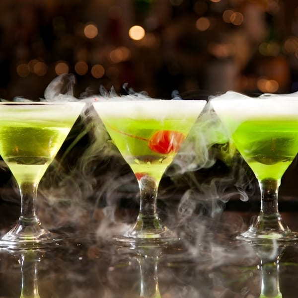 Three smoking green cocktails in martini glasses, one with a cherry, on a dark bar.