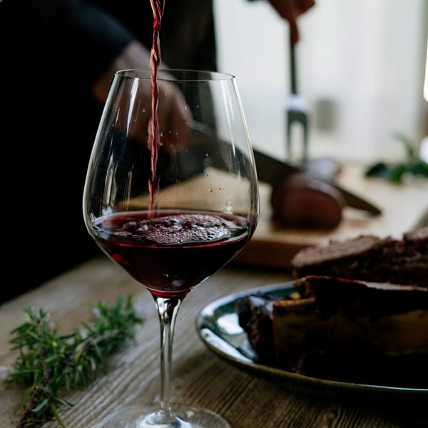 Red wine pouring into a glass next to sliced meat and rosemary on a wooden table.